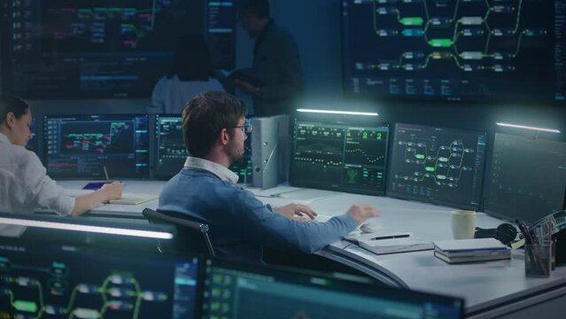 Software engineer works at computer in monitoring room. Technical support analyzes server data late night. Multiple big screens on the wall with displayed real-time analysis charts. Cyber security.