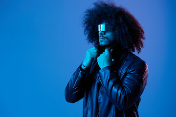 Portrait of fashion man with curly hair with stylish glasses on blue background multinational, colored light, black leather jacket trend, modern concept.