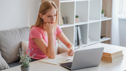 Home study. Computer concentrated. Learning frustration. Tired young girl sitting chin on hand writing notes with pen at table with laptop in light room.