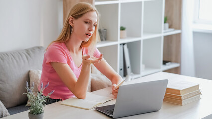 Internet research. Challenging learning. Self-education. Pensive confused young girl sitting with question gesture and pen in hand at table with laptop and notebook in light interior.