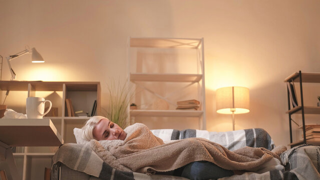 Home rest. Weekend relaxation. Tired woman laying couch under plaid napping day sleeping light shadow room interior.