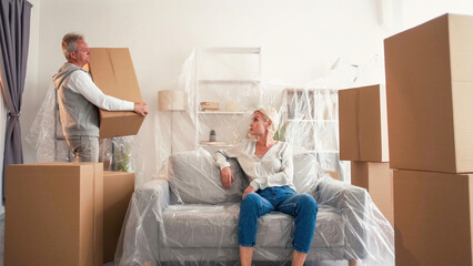 Fototapeta na wymiar Move apartment. Unpacking belongings. Woman sitting sofa in plastic film when husband taking property craft boxes in light room interior.