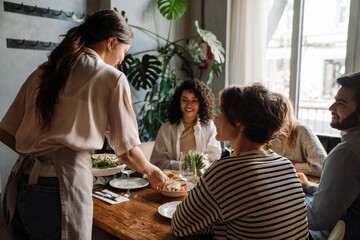 Young waitress serving group of cheerful friends in restaurant