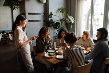 Beautiful waitress serving group of cheerful friends in restaurant