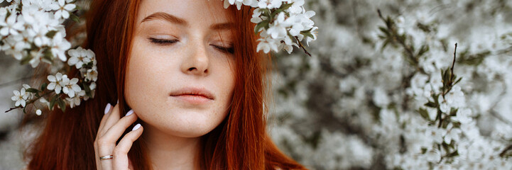 A young beautiful red-haired woman stands near a branching spring apple tree. A girl in a white dress smiles between blossoming apple trees. Banner for website header design with copy space.