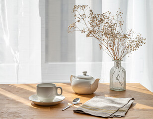 A cup of tea, teapot and dry bouquet flowers on the wooden table of a window with morning sunlight....