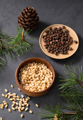 Pine nuts in a bowl and a scattered on a dark background with branches of pine needles and cone. The concept of a natural, organic and healthy superfood and snack.
