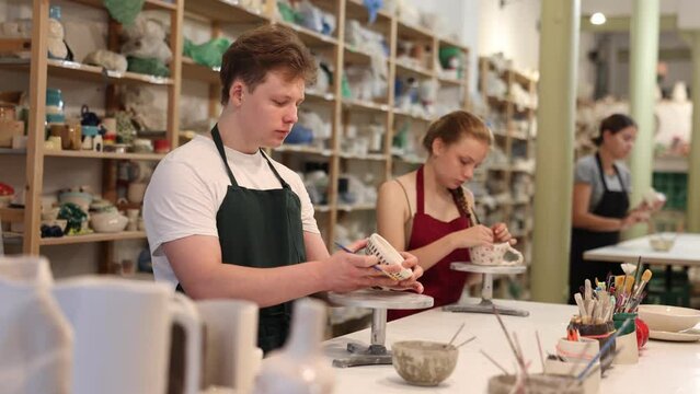 Young woman teacher showing teenage boy and girl how to paint ceramic cup