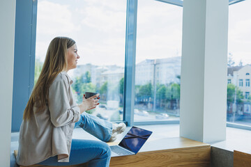 Fototapeta premium Pregnant cheerful woman sitting with laptop, smartphone, coffee cup, looking at laptop and taking notes while sitting in cafe. A woman works remotely on a laptop while on maternity leave