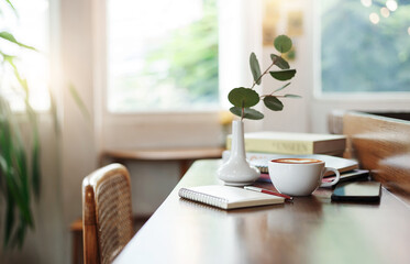 Coffee cup on wooden counter and smart phone, note book put on digital tablet with ceramic vase and small tree, coffee break after work in cafe with reading book in the background
