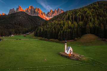 Val Di Funes, Dolomites, Italy - Aerial view of the beautiful St. Johann Church (Chiesetta di San Giovanni in Ranui) at South Tyrol with Italian Dolomites in warm sunset colors at background at summer