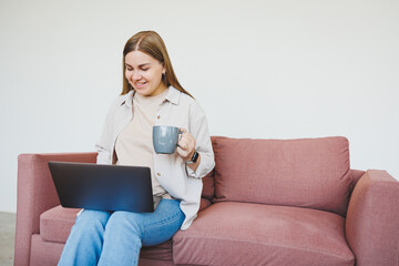 Smiling female freelancer in casual clothes sitting in comfortable sofa in modern cafeteria at table with laptop and phone, talking on mobile phone. Remote work