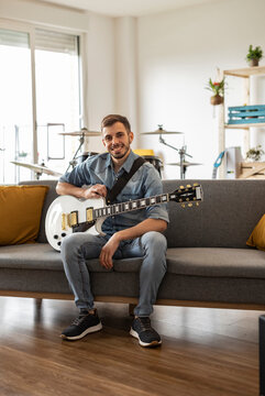 Happy young guitarist sitting with guitar on sofa at home