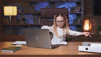Vibrant blonde kid chilling out and listening to drive music on wireless laptop in coworking space. Energetic little girl moving rhythmically hands while resting on comfortable office chair.