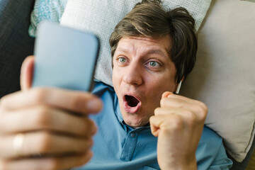 Excited man holding smart phone lying down on bed at home