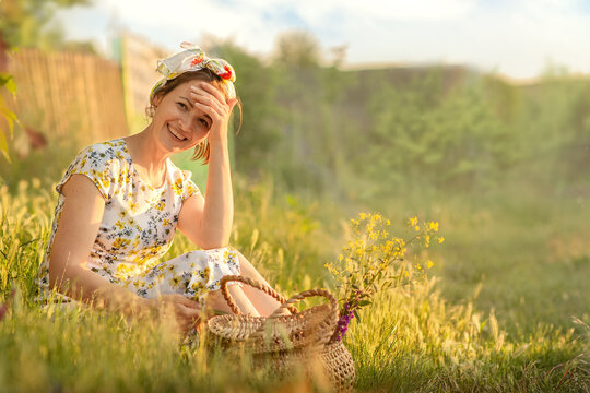a young woman at sunset is sitting on the side of the road in the grass with a basket of flowers,the sunset beautifully illuminates the girl,she is in a very good mood,laughing merrily into the camera