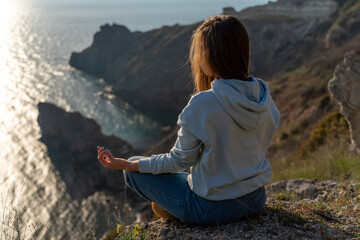 Naklejka na ściany i meble Woman tourist enjoying the sunset over the sea mountain landscape. Sits outdoors on a rock above the sea. She is wearing jeans and a blue hoodie. Healthy lifestyle, harmony and meditation