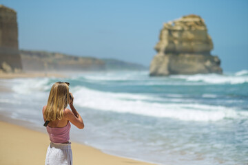 Young beautiful woman walking on the beach and taking photos at Twelve Apostles rock formations at the great ocean road in sunny weather with a blue sky, Victoria, Australia 