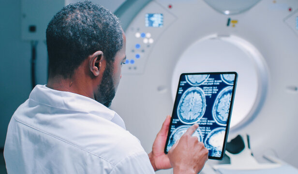 African American doctor examine scan attentively. Medical worker shooted from back. Male doctor examines MRI scan. Multicultural doctor is scrolling screen of tablet with scans of brain.