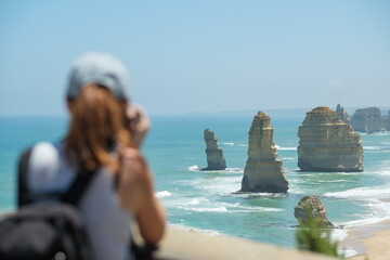 Young beautiful woman taking photos at Twelve Apostles rock formations at the great ocean road in sunny weather with a blue sky, Victoria, Australia, focused on the background, woman is blurred