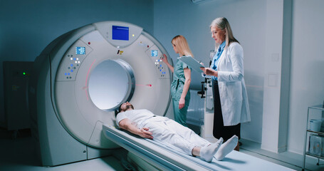 Young man lies down in MRI capsule. Chief head doctor in dressing gown hold folder and explains to patient what to do. Nurse monitors process of examiming. Man undergoes magnetic resonance imaging.