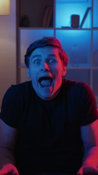 Vertical video. Grimacing man. Meme expression. Silly behavior. Foolish guy making nuts scare face sitting sofa in dark neon light interior looped.