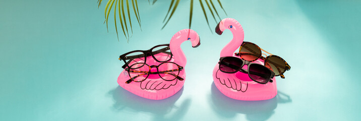 Optic store advertisement banner. Clear Eyeglasses Glasses with plastic Frame and trendy sunglasses on inflatable flamingo on blue background with palm leaves. Copy space. Optic store web line