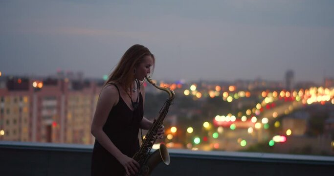 A young woman plays the saxophone while standing on the roof of a building in the city center late at night. Romantic music on the rooftop - 25 fps 4k footage.