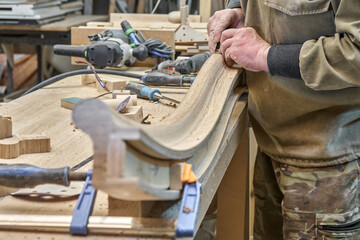 Carpenter sands bending wooden railing with sandpaper in workshop closeup. Senior master makes detail of spiral staircase for home interior