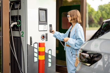 Young woman going to charge electric car on vehicle public power station, scanning QR code with...