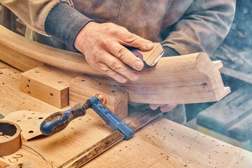 Carpenter sands bending wooden railing with sandpaper in workshop closeup. Senior master makes detail of spiral staircase for home interior