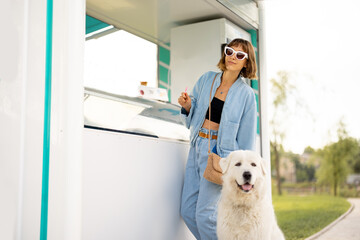 Young woman choosing ice cream to buy at beautiful shop while walking with her dog in a park....