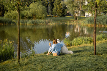 Young woman eats ice cream while sitting with her cute white dog near the lake in park. Wide view. Friendship with pets and spending leisure time together