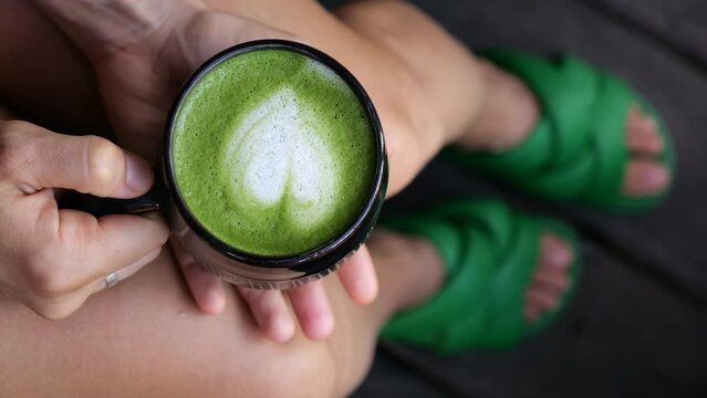 A woman sitting at home holds a cup of hot matcha latte in her hands, relaxation for the whole day from the hustle and bustle with a hot drink. Home concept A cozy moment with a cup of hot matcha tea.