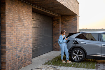 Young woman using smart phone while charging her electric car near garage of her house. Concept of modern lifestyle and sustainability - 616937698