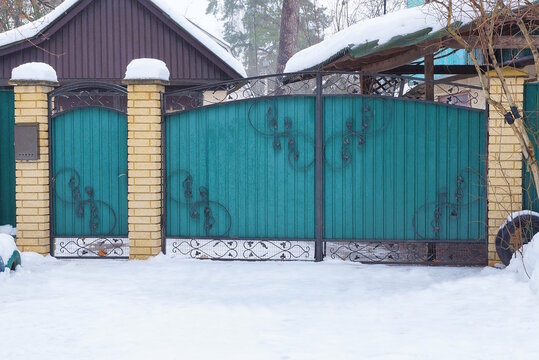 a large closed metal green gate and a closed iron door with a black forged pattern in a snowdrift of white snow on a winter rural street