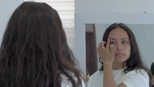 young woman looks at herself in the mirror, eyecontact, checking her face, pretty look, natural beauty, dark hair,  female face medium wide reflection, bathroom mirrors, make-up eyebrow, lashes check.