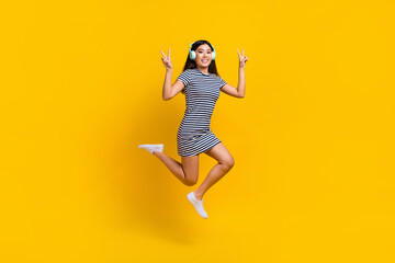 Fototapeta na wymiar Full length photo of cheerful pretty lady wear striped dress earphones jumping showing v-signs isolated yellow color background