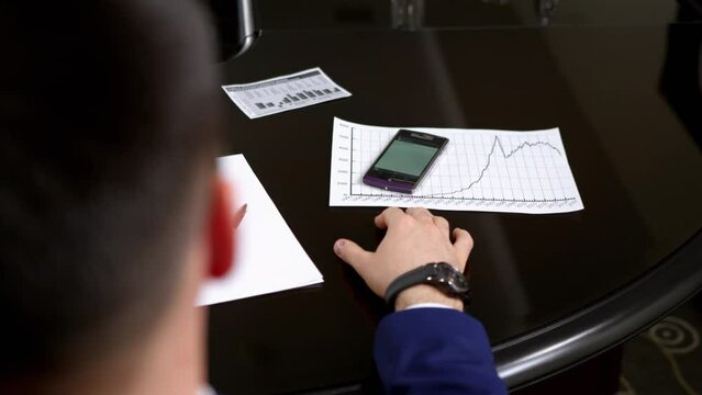 Businessman looks at and takes ringing cell phone in meeting crop body back view. Purple mobile phone stands on black office table