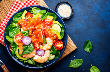 Fresh salad for keto diet with salmon, avocado, spinach, cucumber, shrimps, tomato, cashew, sesame seeds. Low-carbohydrate lunch rich in healthy fats. Blue stone table background, top view