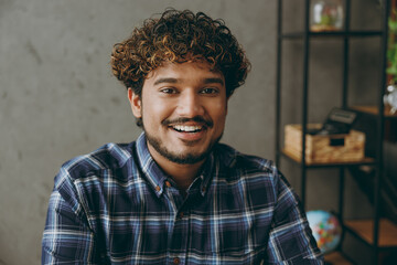 Close up successful cheerful satisfied positive fun cool smiling happy smart employee business Indian man he wearing casual blue checkered shirt looking camera sitting working at office desk indoors.