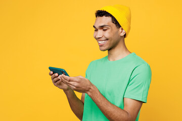 Young man of African American ethnicity he wear casual clothes green t-shirt hat use play racing app on mobile cell phone hold gadget smartphone for pc video games isolated on plain yellow background