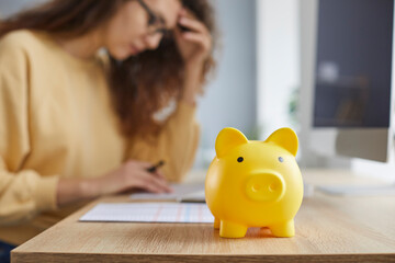 Yellow piggy bank on the table with a young businesswoman accountant in background working at home or at office with documents, tables and charts making calculations. Investment and savings concept.
