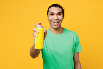 Young man of African American ethnicity he wears casual clothes green t-shirt hat man using spray bottle for painting graffiti isolated on plain yellow background studio portrait. Lifestyle concept.