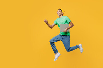 Full body young IT man of African American ethnicity wearing casual clothes green t-shirt hat jump high hold closed laptop pc computer run fast isolated on plain yellow background. Lifestyle concept.