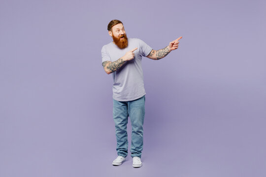 Full body young redhead bearded man wear violet t-shirt casual clothes point index finger aside indicate on workspace area copy space mock up isolated on plain pastel light purple background studio.
