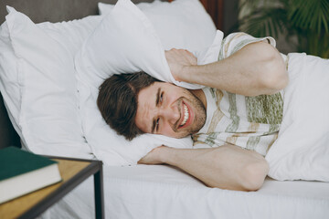 Young sad tired annoyed man wearing casual clothes white t-shirt pajama lying in bed cover ears...
