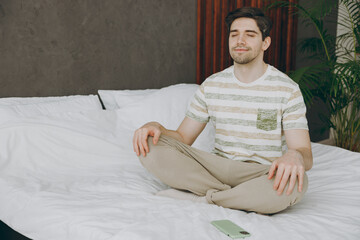 Young spiritual man wearing casual clothes t-shirt pajama sitting on bed meditating try to calm...