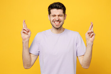 Young man wear light purple t-shirt casual clothes waiting for special moment, keeping fingers crossed, making wish, eyes closed isolated on plain yellow background studio portrait. Lifestyle concept.