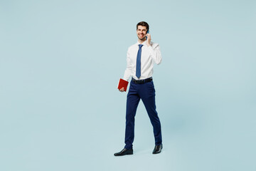 Full body young employee IT business man corporate lawyer wear classic formal shirt tie work in office talk speak on mobile cell phone hold book isolated on plain pastel light blue background studio.